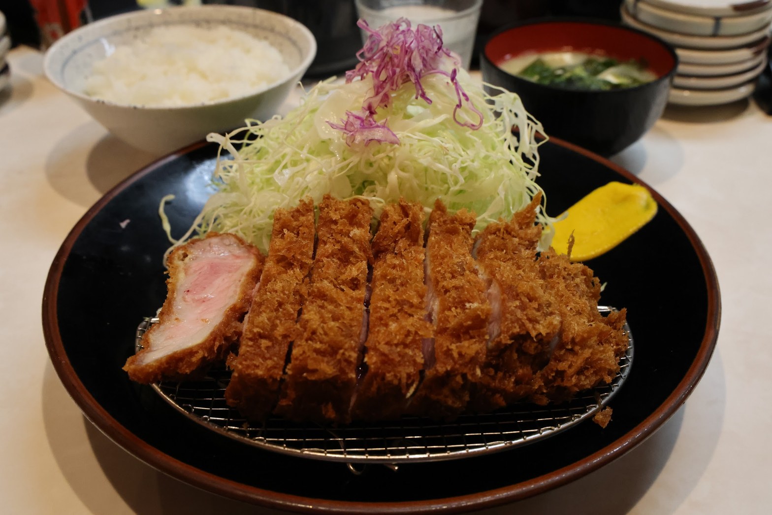 黒い丸皿の上の網に乗った厚切りとんかつの断面と、盛り上がったキャベツの千切り、奥に白飯と味噌汁