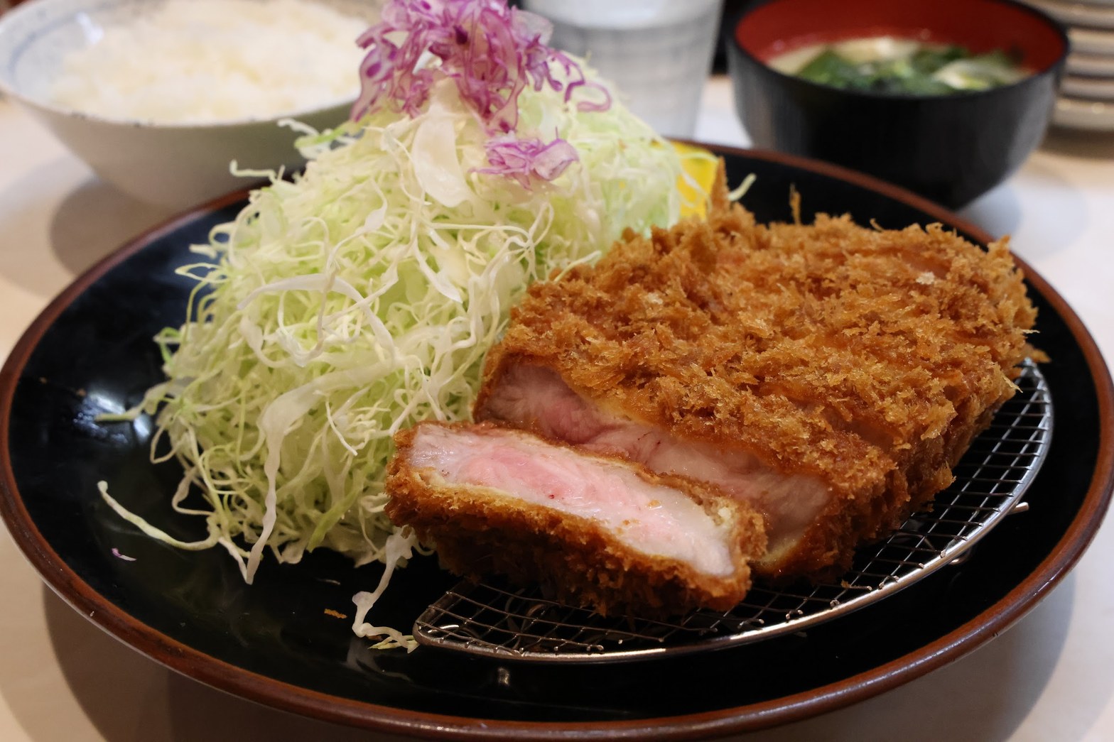 黒い丸皿に盛られた揚げたてとんかつの断面とキャベツの千切り、奥に白飯と味噌汁の椀
