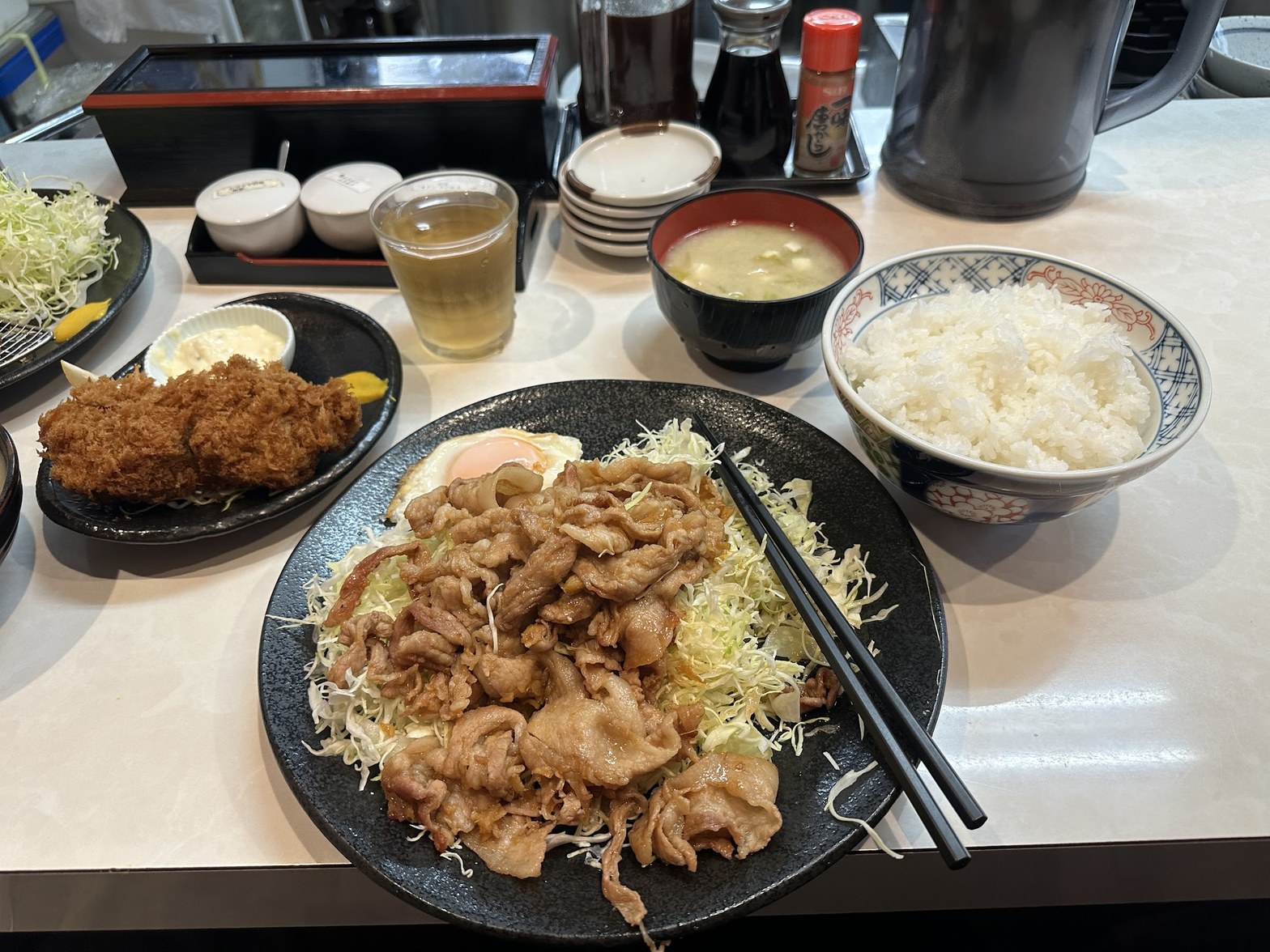 コの字カウンターに並ぶ生姜焼きと小皿のとんかつ、白飯の茶碗と味噌汁、卓上の調味料